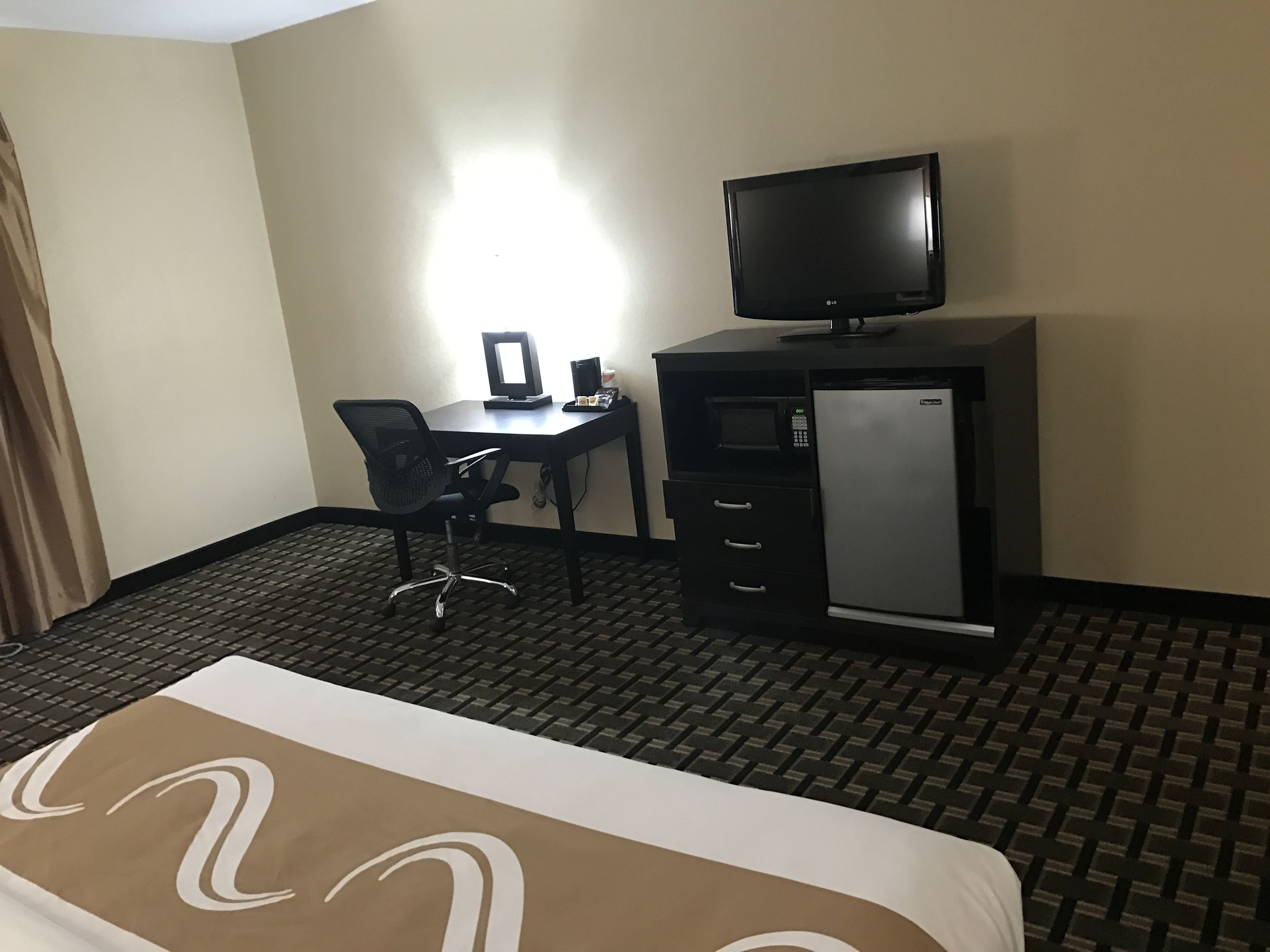 A hotel room with a bed, a desk with a computer, a flat-screen TV, and a mini-fridge.