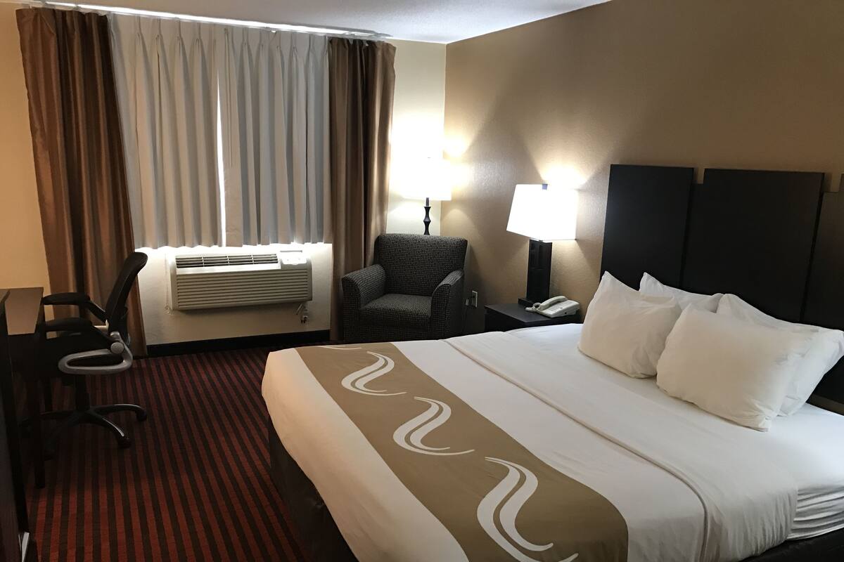 A hotel room with a large bed, a chair, an air conditioning unit, a lamp, and a telephone on the nightstand.