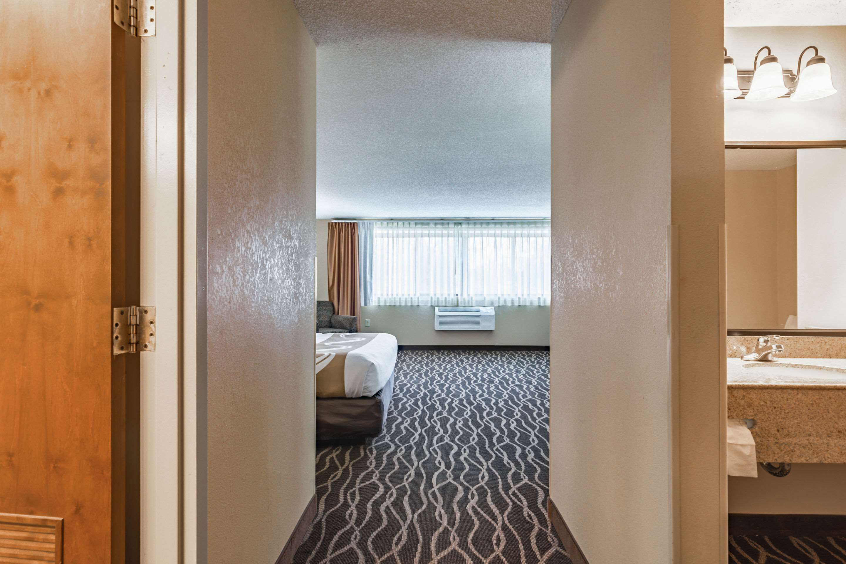 Una camera d'albergo con tappeto a motivi, un letto, una finestra con tende, un condizionatore d'aria e un bagno con lavandino e specchio.