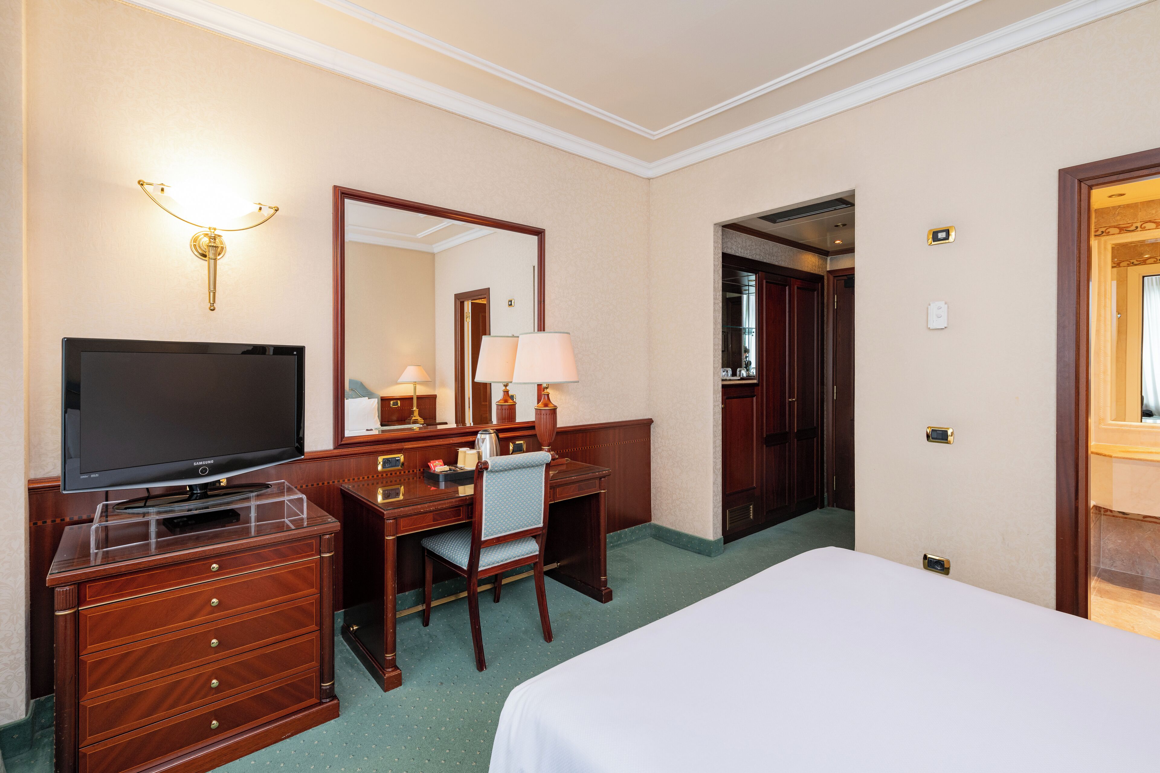 A hotel room with a bed, a desk with a television, a chair, and a desk lamp.