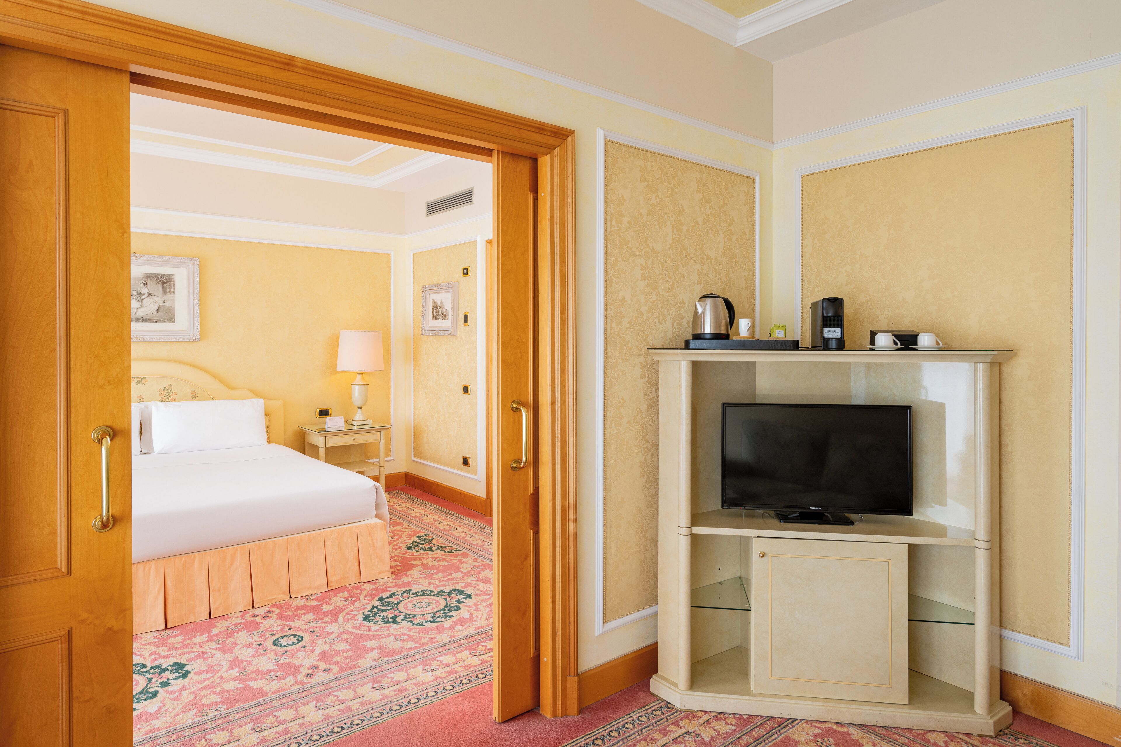 A hotel room with a bed, a television on a stand, a nightstand with a lamp, and a carpeted floor.