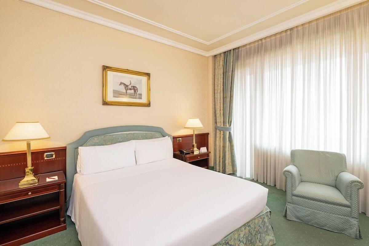Une chambre d’hôtel avec un lit, deux tables de chevet avec des lampes, une chaise, un tableau encadré au mur et une fenêtre avec des rideaux.