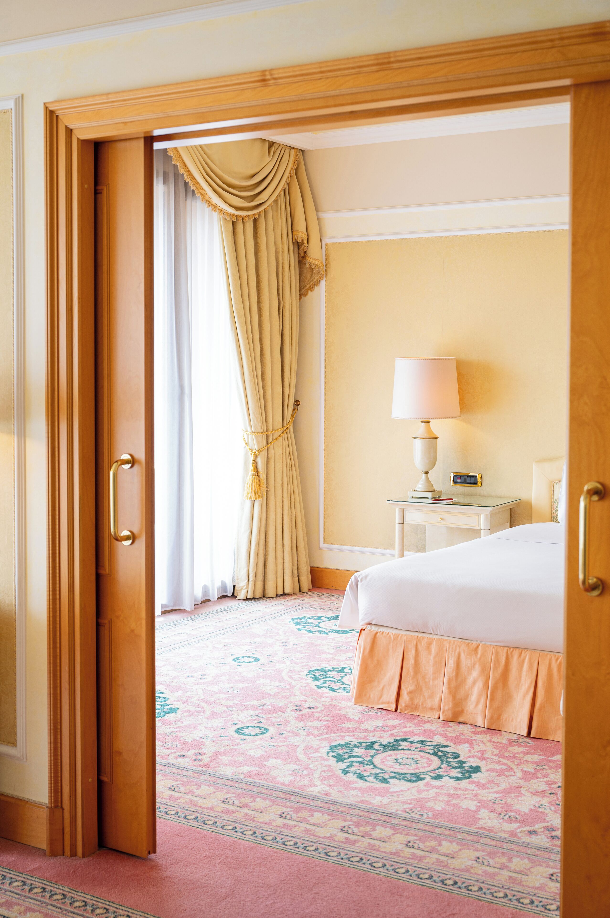 A hotel room with a bed, a nightstand with a lamp, a clock, and a patterned carpet.