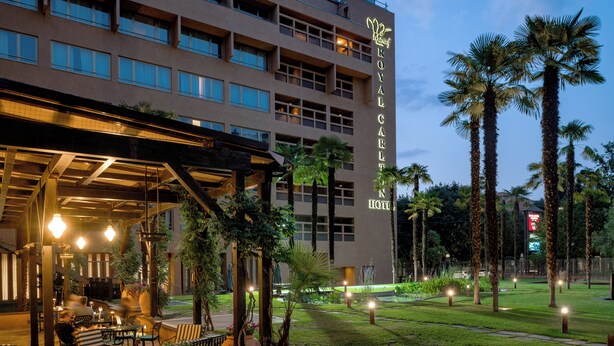 A hotel with a modern facade, outdoor seating area, and palm trees. - Royal Hotel Carlton (Bologna)