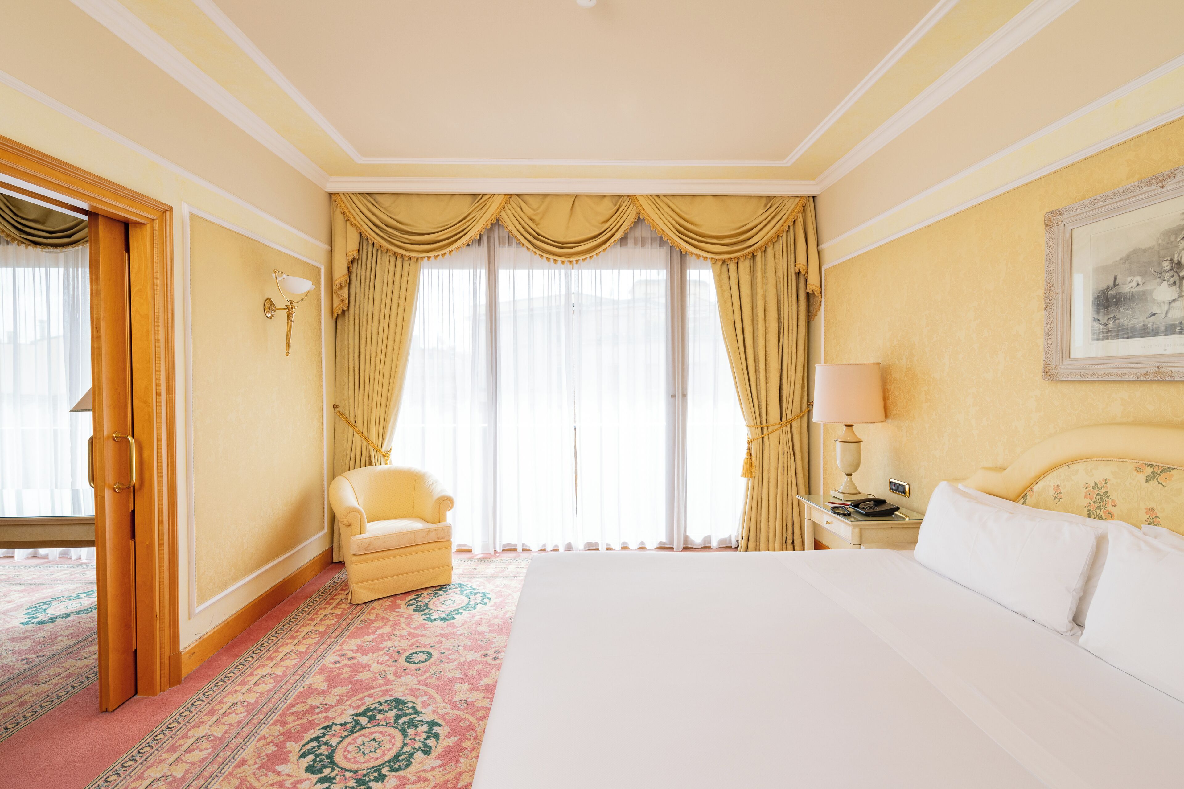 A hotel room with a large bed, a chair, a nightstand with a lamp, and a painting on the wall.