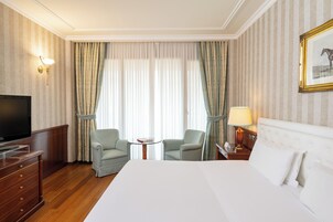 A hotel room with a large bed, two armchairs, a small table, a TV, and curtains.