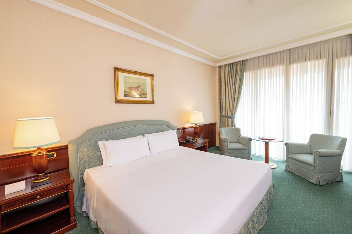 Une chambre d’hôtel avec un grand lit, deux tables de chevet avec des lampes, une chaise, une petite table et une fenêtre avec des rideaux.