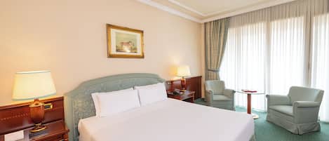 A hotel room with a large bed, two bedside tables with lamps, a chair, a small table, and a window with curtains.