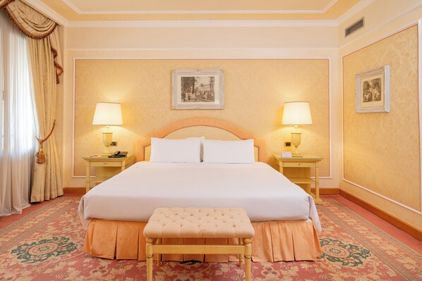 Una habitación de hotel con una cama grande, dos mesitas de noche con lámparas, un banco y una alfombra estampada.