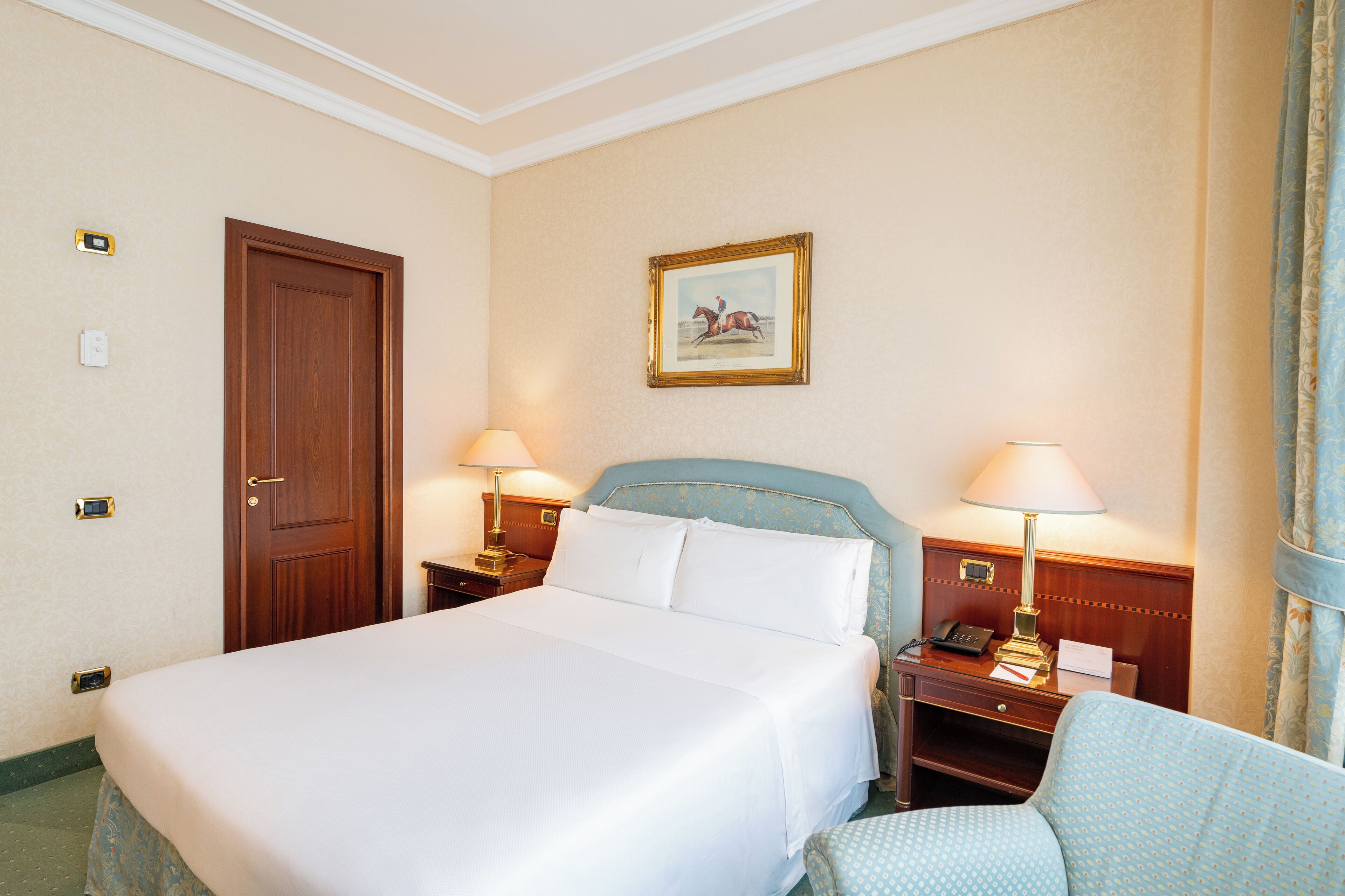 A hotel room with a large bed, two bedside tables with lamps, a wooden door, a framed picture on the wall, and a window with curtains.
