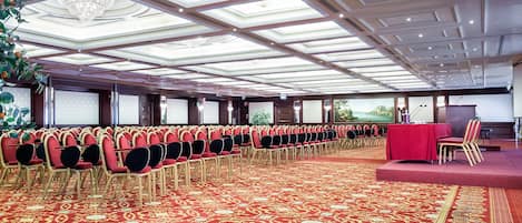 A conference room with rows of chairs, a stage with a podium, and a large chandelier.