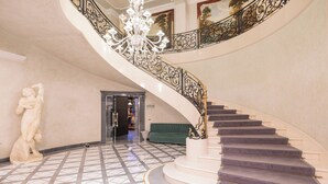 A grand staircase with ornate black railings and a chandelier, leading to a room with a green sofa and a statue. - Royal Hotel Carlton (Bologna)