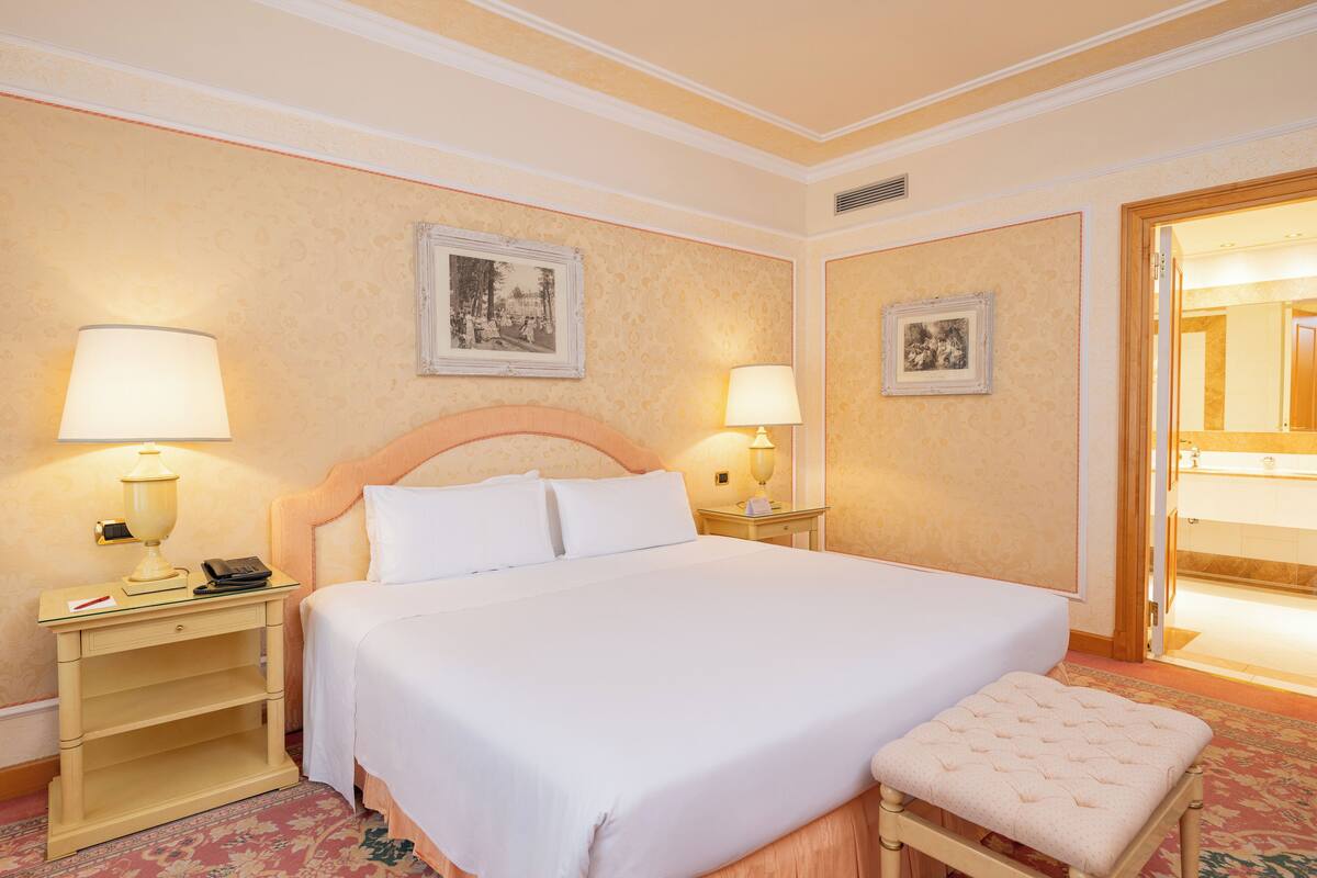 Une chambre d’hôtel avec un grand lit, deux tables de chevet avec des lampes, un petit banc et deux tableaux encadrés accrochés aux murs.
