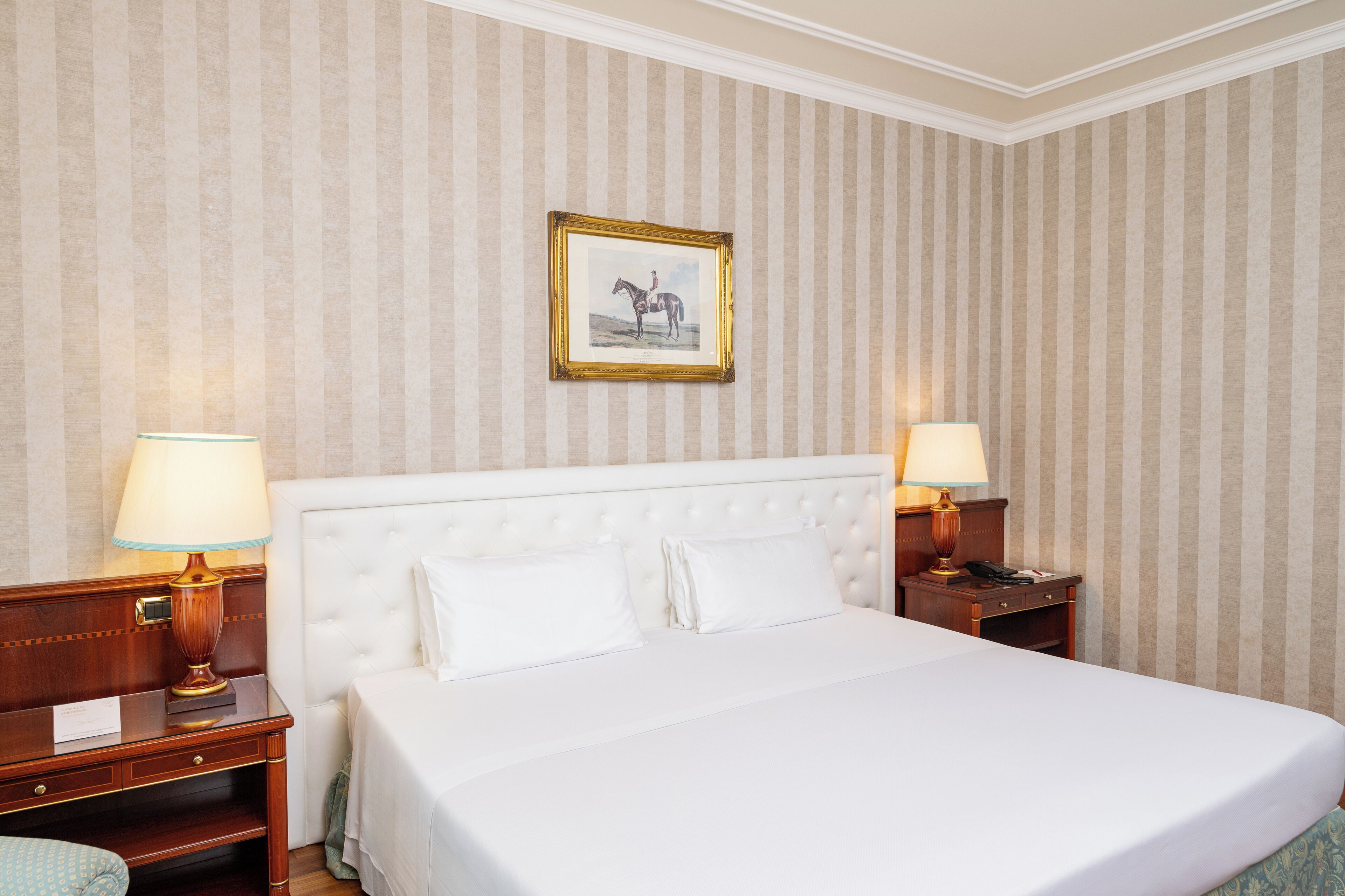 Une chambre d’hôtel avec un grand lit, deux tables de chevet avec des lampes, un tableau encadré au mur et un papier peint à rayures.