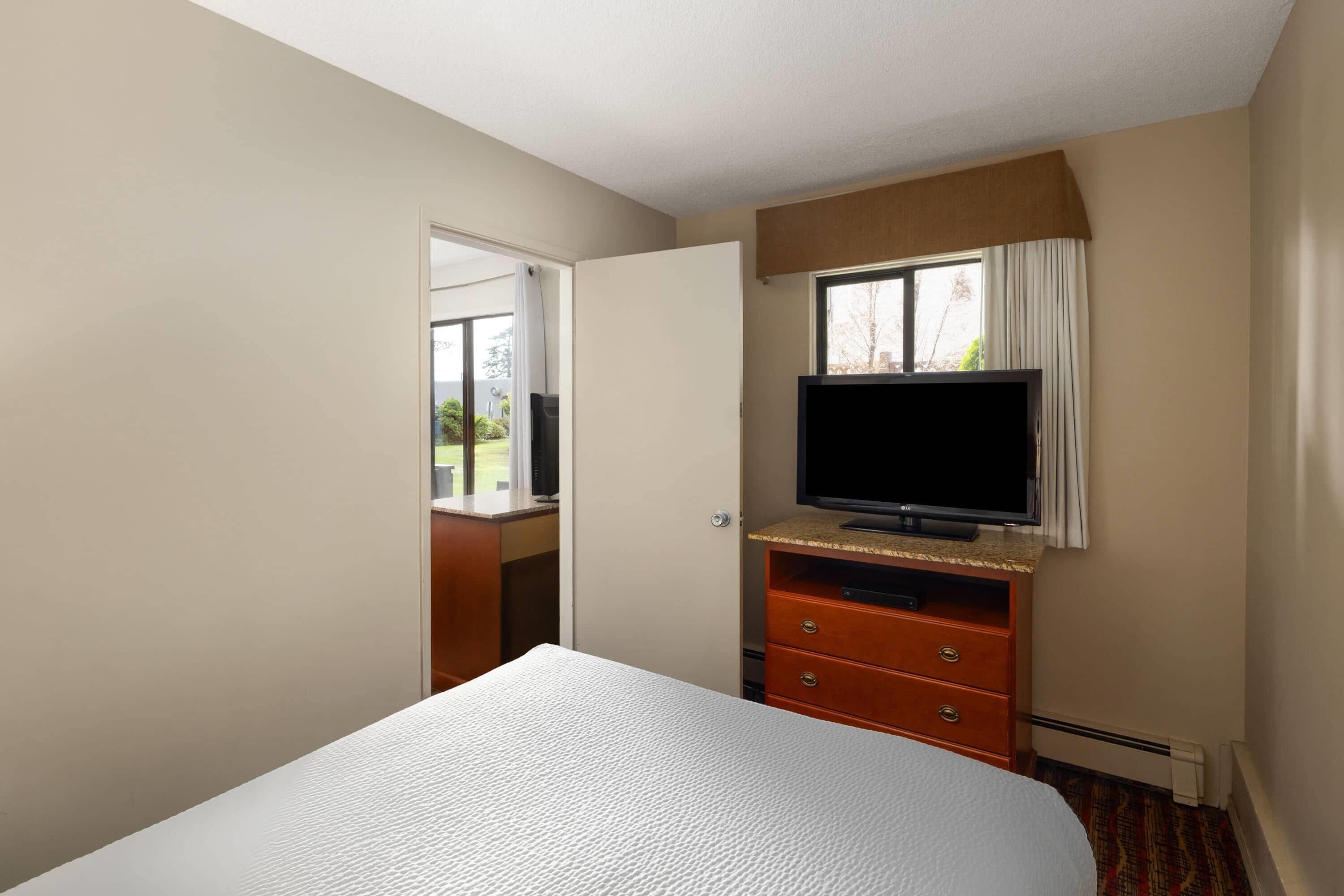 Quarto de hotel com uma cama, uma televisão sobre um suporte de madeira, uma janela com cortinas e vista para o exterior.