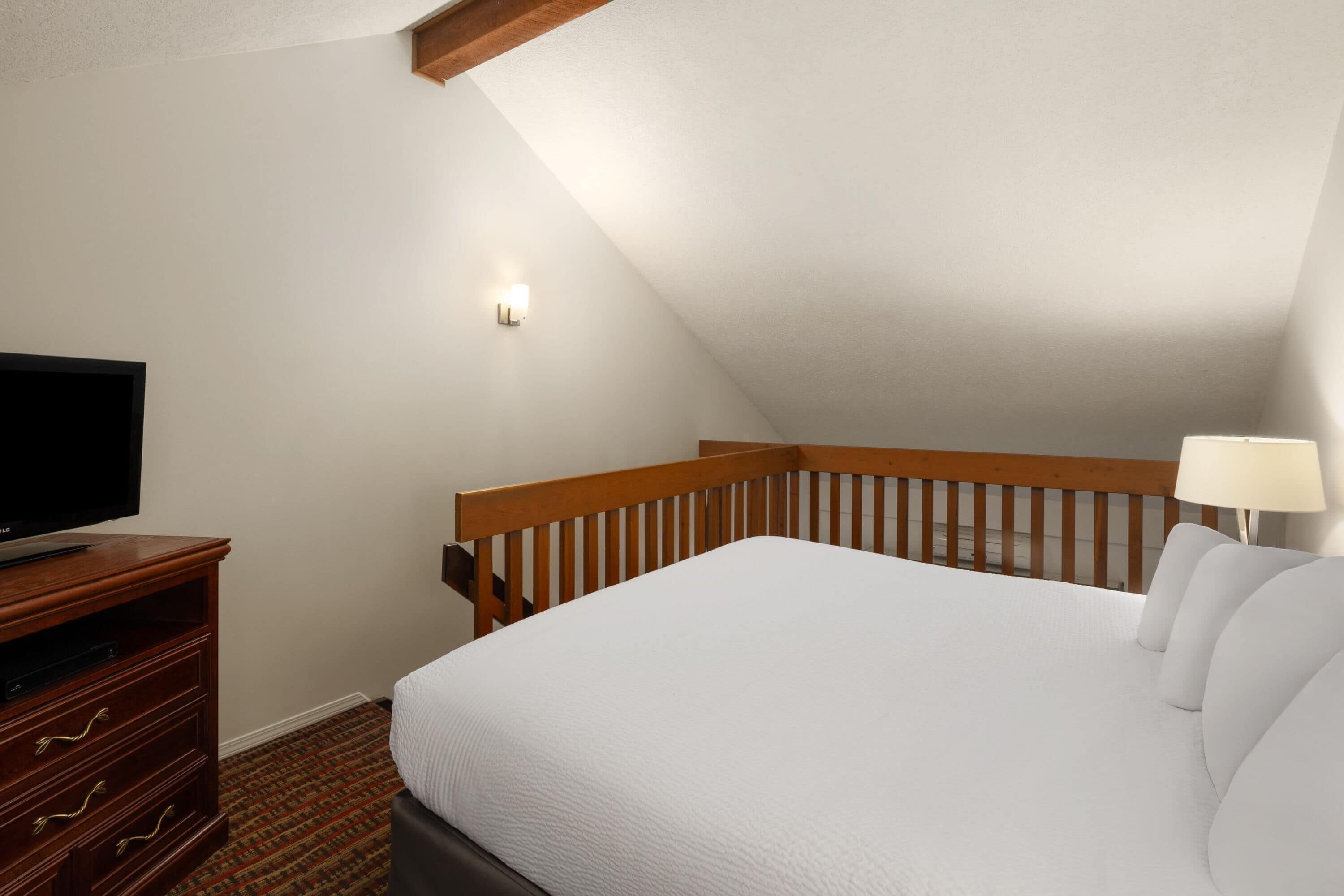 A bedroom with a bed, a TV on a wooden stand, a nightstand, a lamp, and a sloped ceiling.