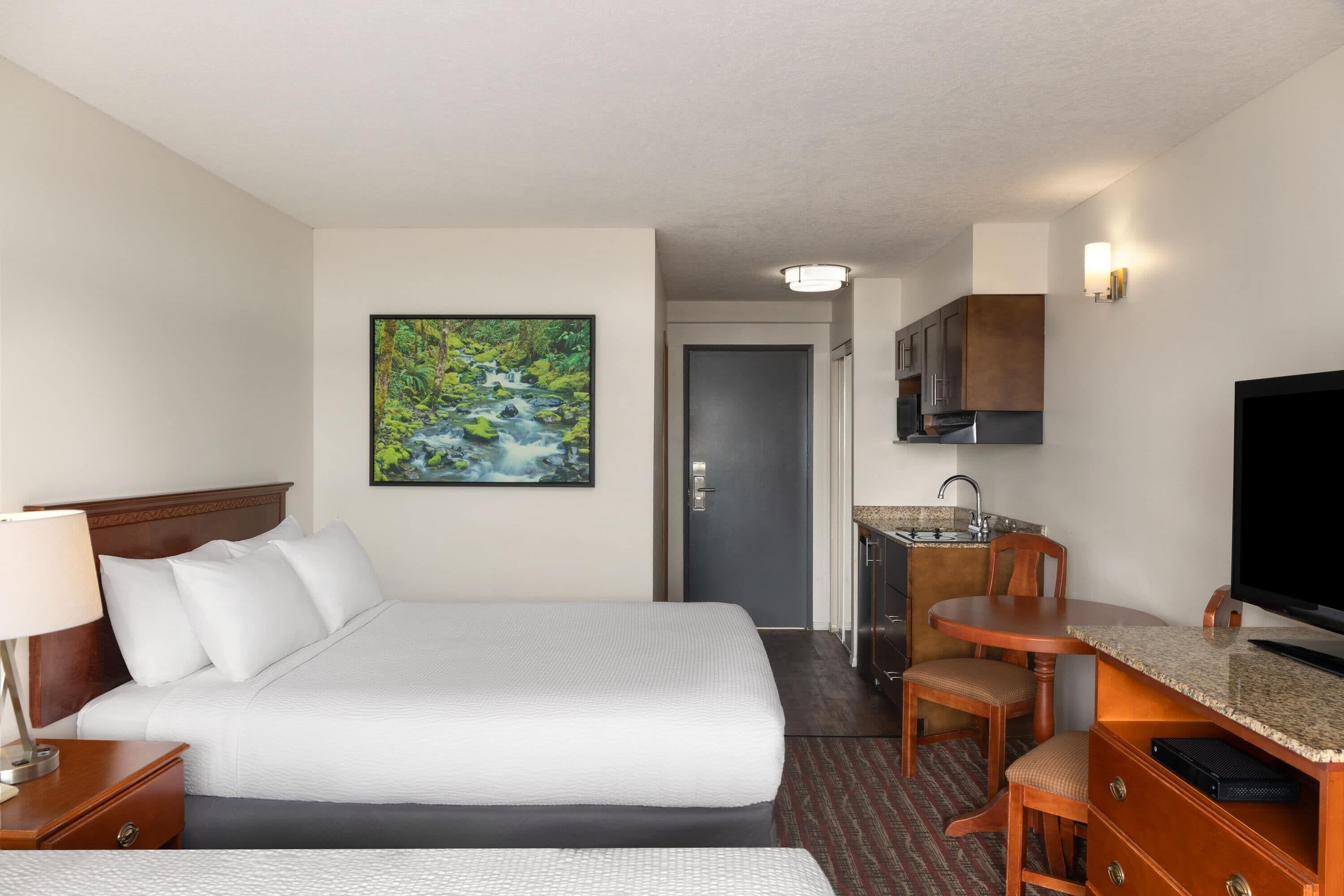 A hotel room with a bed, a desk with a chair, a television, and a painting of a waterfall.