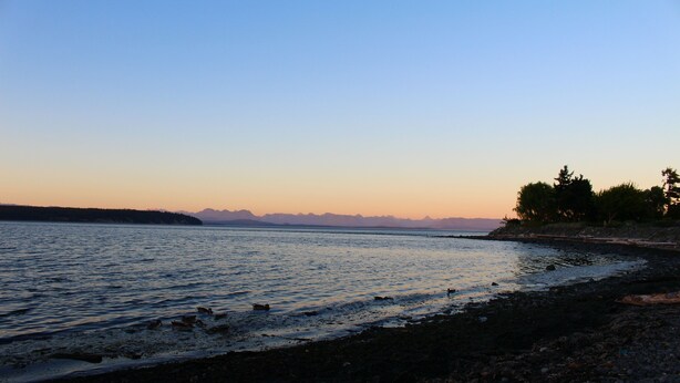 Ein Strand mit ruhigem Wasser, felsigem Ufer und Bergen im Hintergrund.