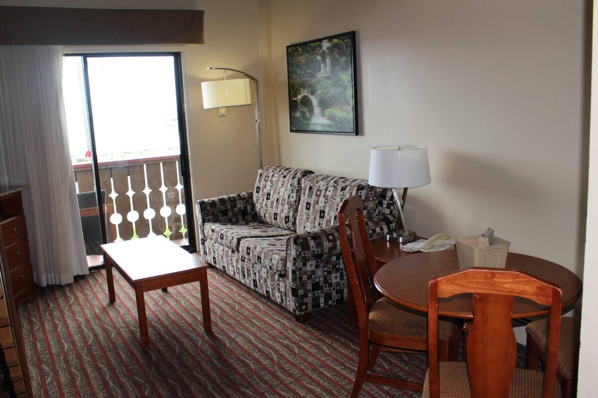 Ein Hotelzimmer mit einer gemusterten Couch, einem Holztisch, einem Stuhl, einer Lampe und einem Bild an der Wand.