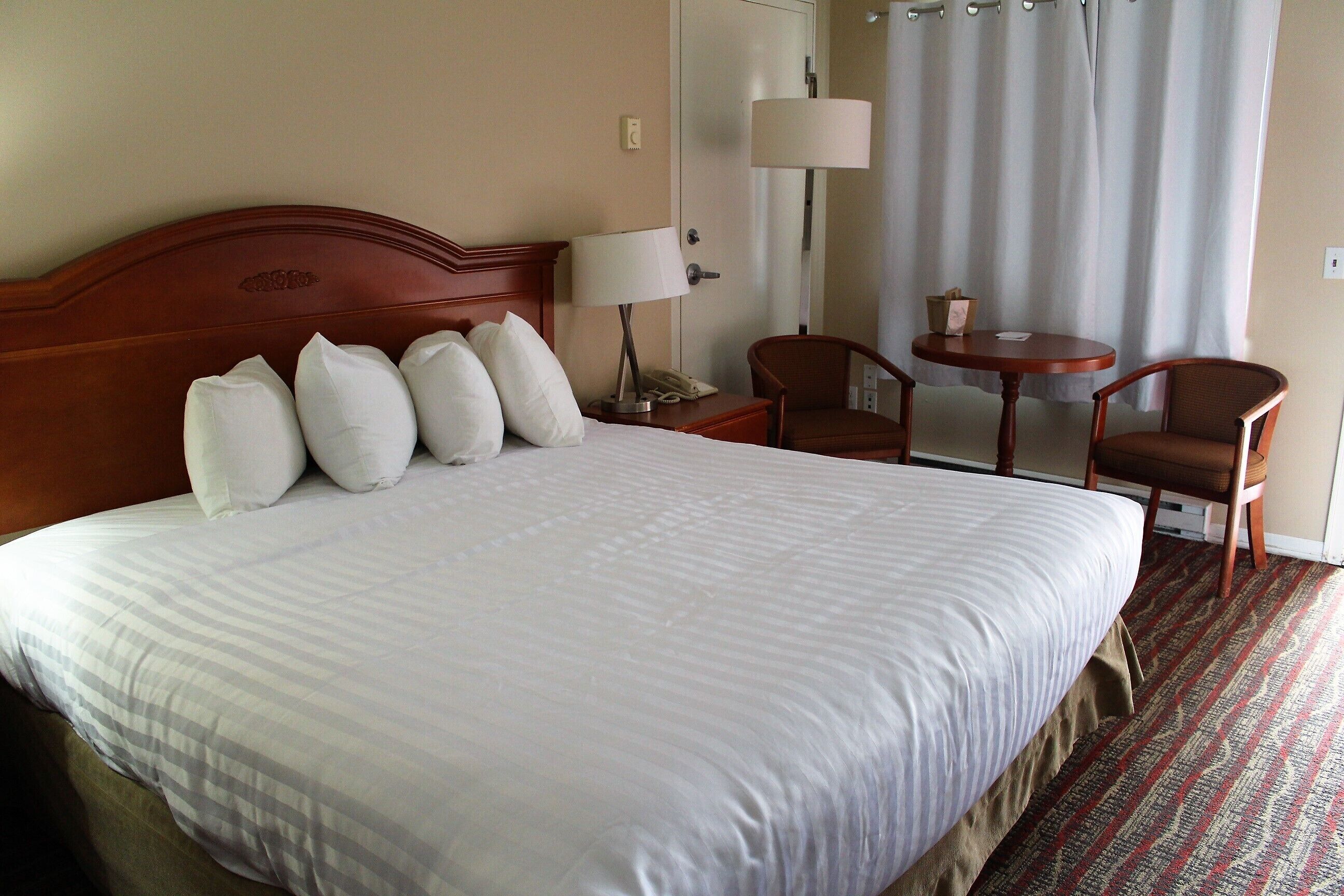 A hotel room with a large bed, a wooden headboard, a bedside table with a lamp, two chairs, and a small table.