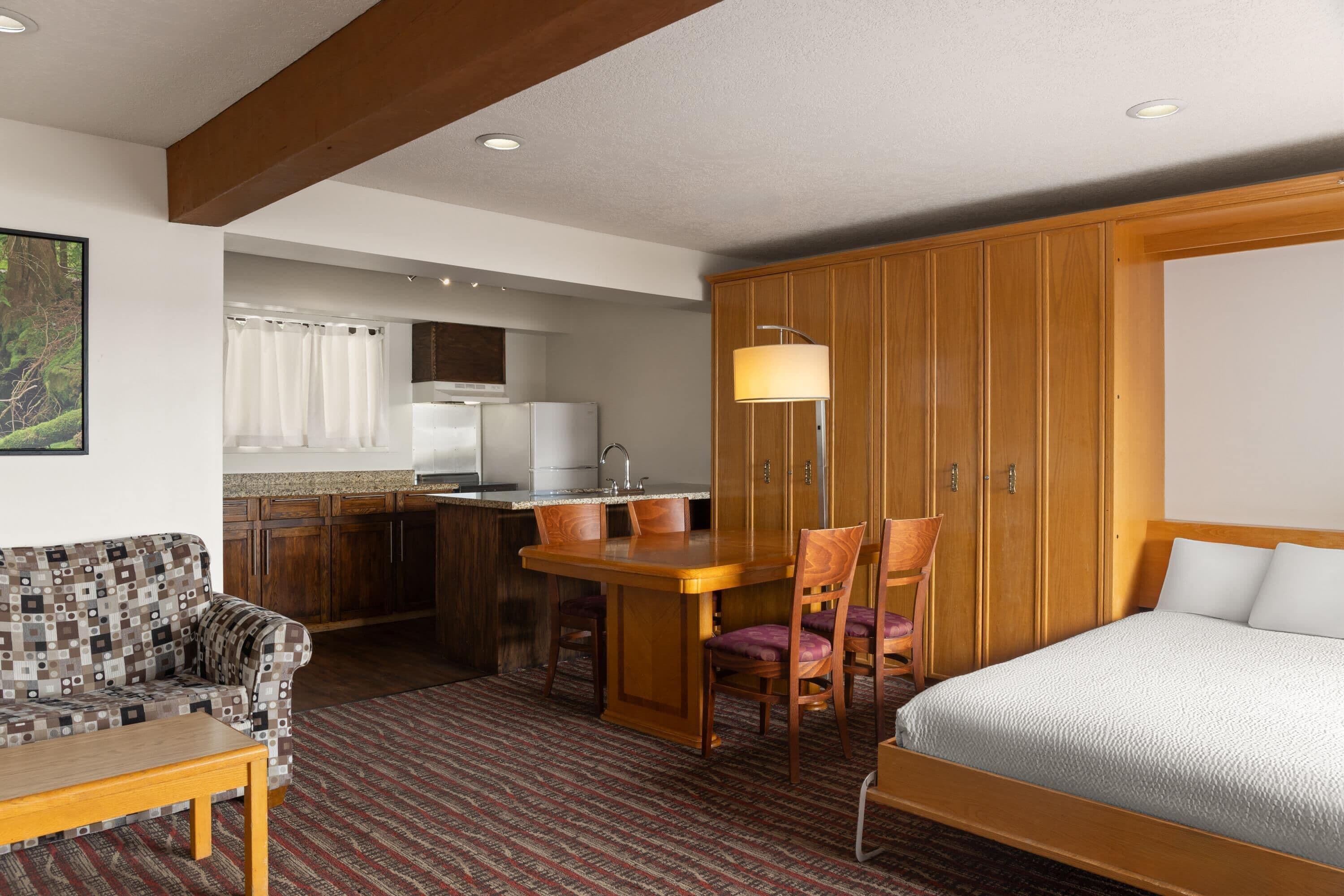 A hotel room with a bed, a wooden desk with chairs, a kitchenette, and a patterned sofa.