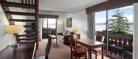 Habitación de hotel con zona de comedor, sofá, escalera y un amplio ventanal con vistas al exterior.