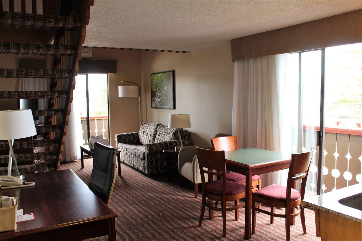 Ein Hotelzimmer mit Esstisch, zwei Stühlen, einer Couch, einem Schreibtisch mit Lampe, einem Telefon und einem Bücherregal.