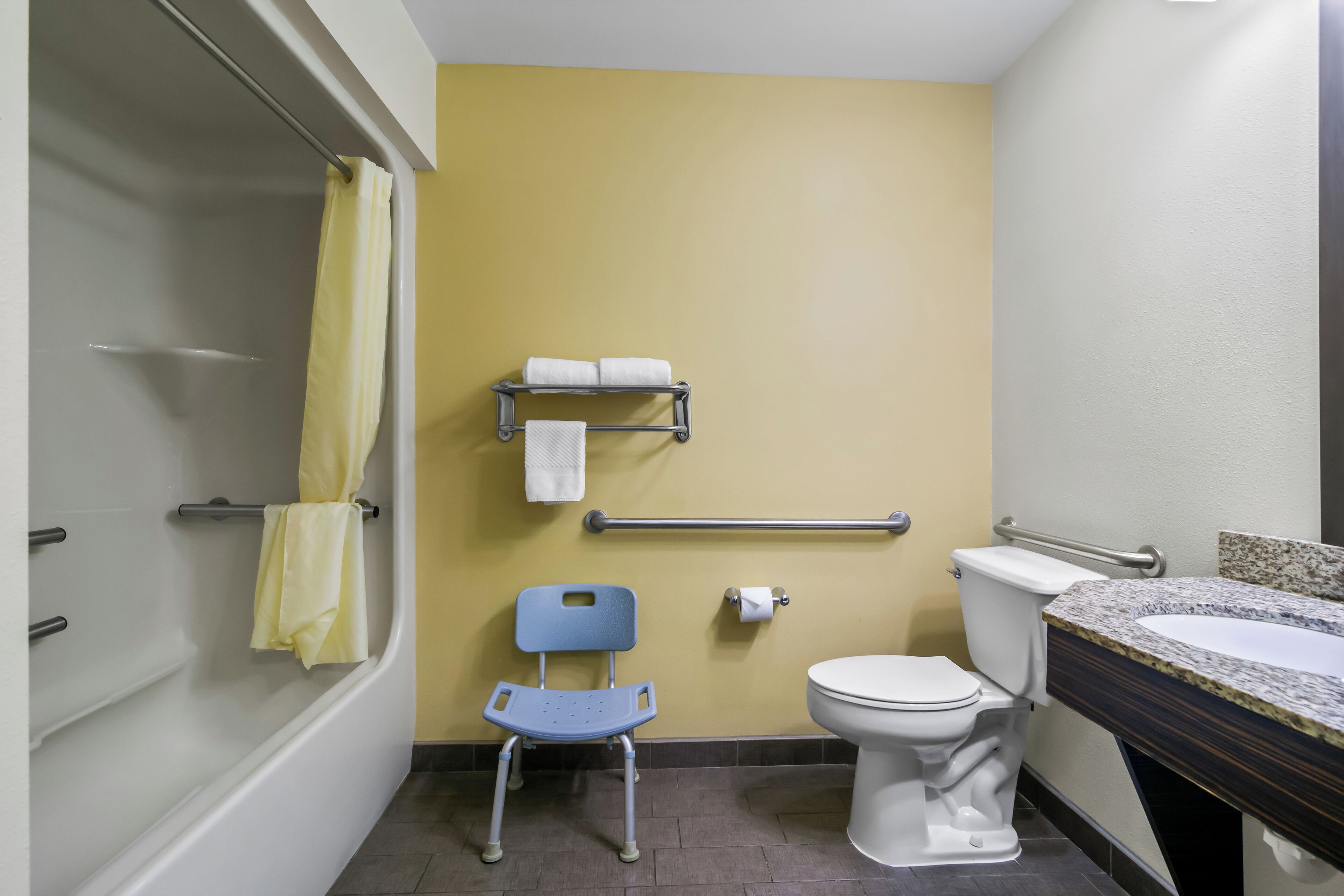 A bathroom with a shower, a toilet, a sink, a grab bar, a chair, and a towel rack.