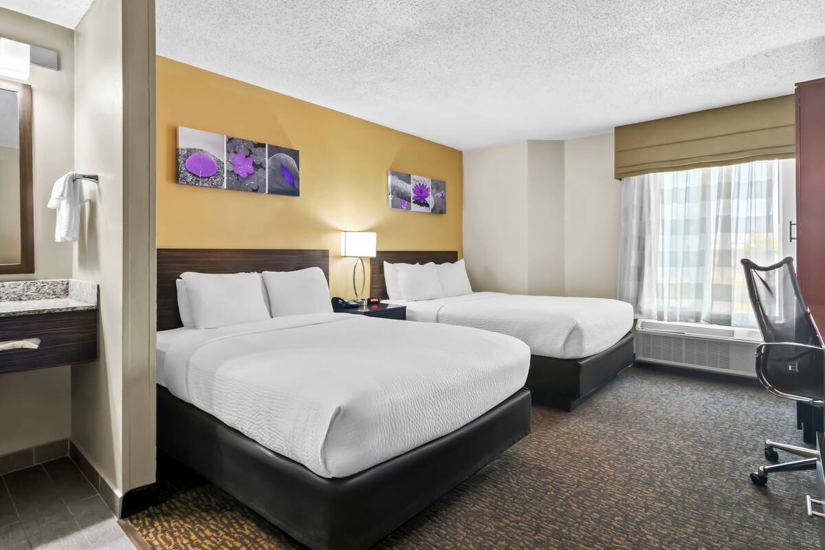 A hotel room with two beds, a desk, a chair, a lamp, and artwork on the wall.
