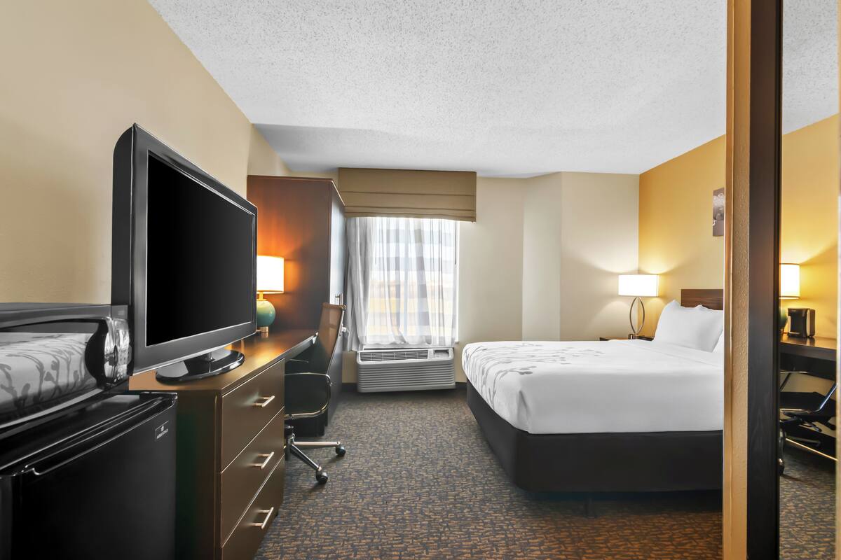 A hotel room with a bed, a desk with a computer, a television, and a window with curtains.