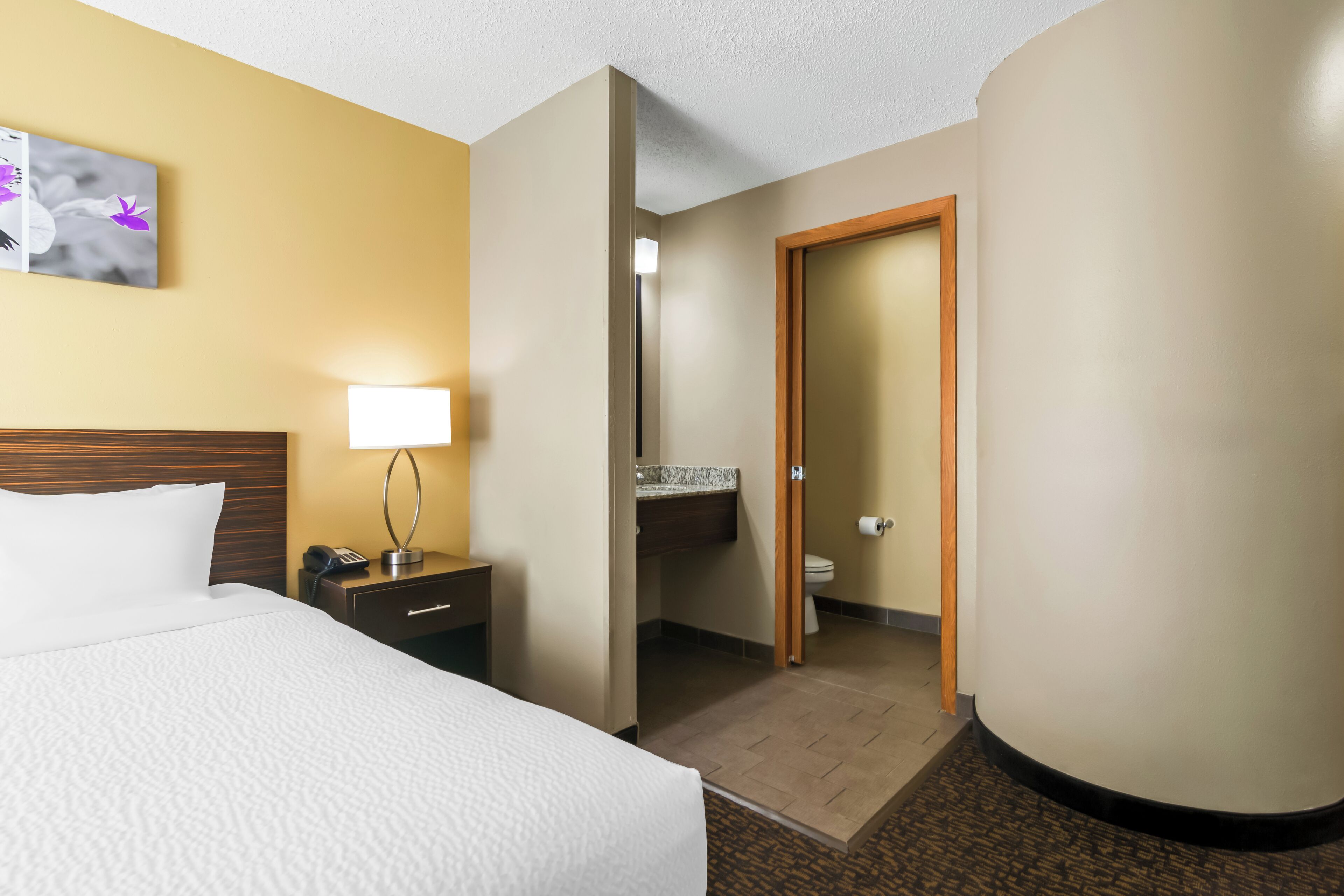 A hotel room with a bed, bedside table, lamp, and a bathroom visible through an open door.