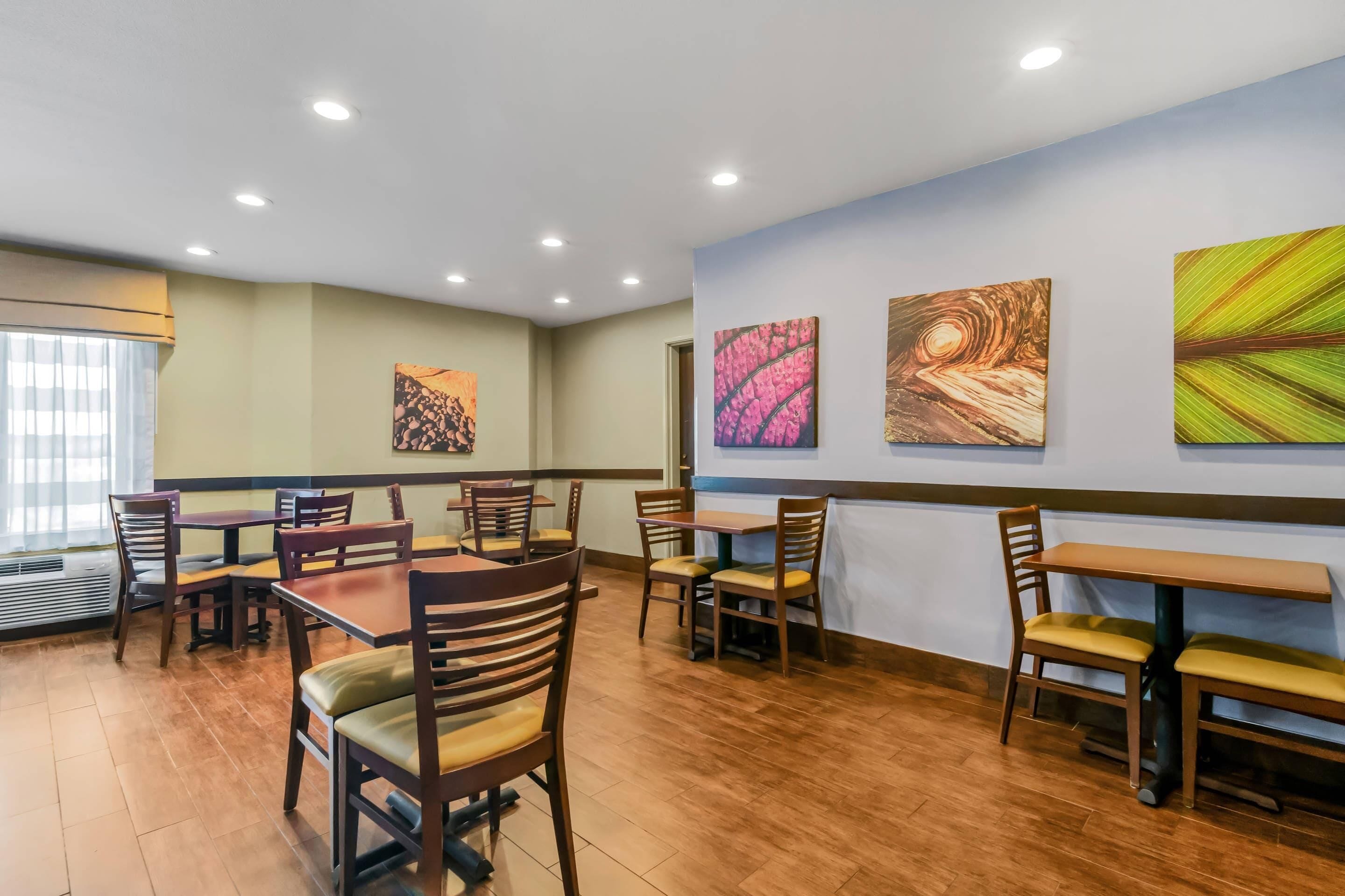 Un comedor con mesas y sillas de madera, un ventanal con persianas y tres cuadros abstractos en la pared.