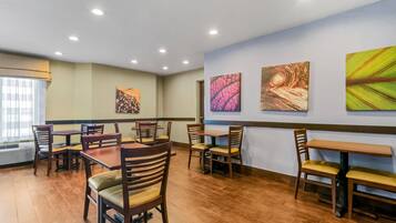 Un comedor con mesas y sillas de madera, un ventanal con persianas y tres cuadros abstractos en la pared.