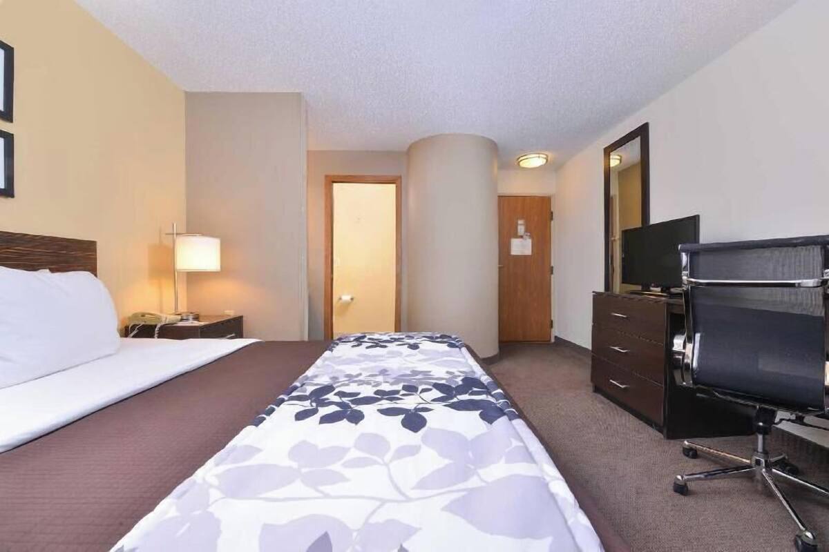 A hotel room with a bed, a desk with a lamp, a TV, and a chair.
