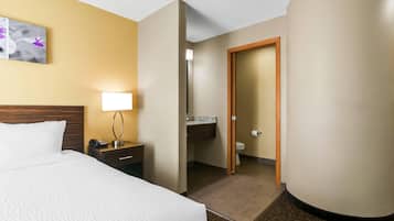 Una habitación de hotel con cama, mesita de noche, lámpara y un baño visible a través de una puerta abierta.