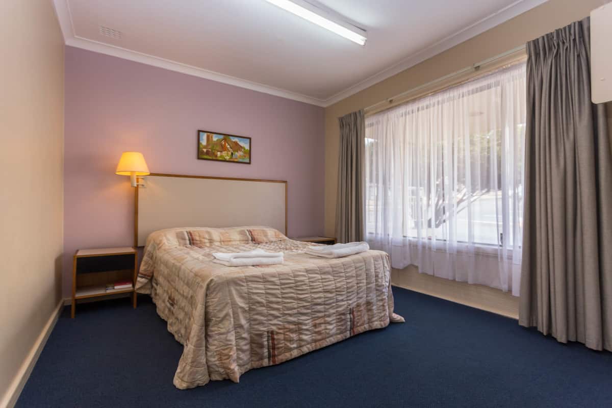 A hotel room with a large bed, a nightstand, a lamp, a painting on the wall, and a window with curtains.