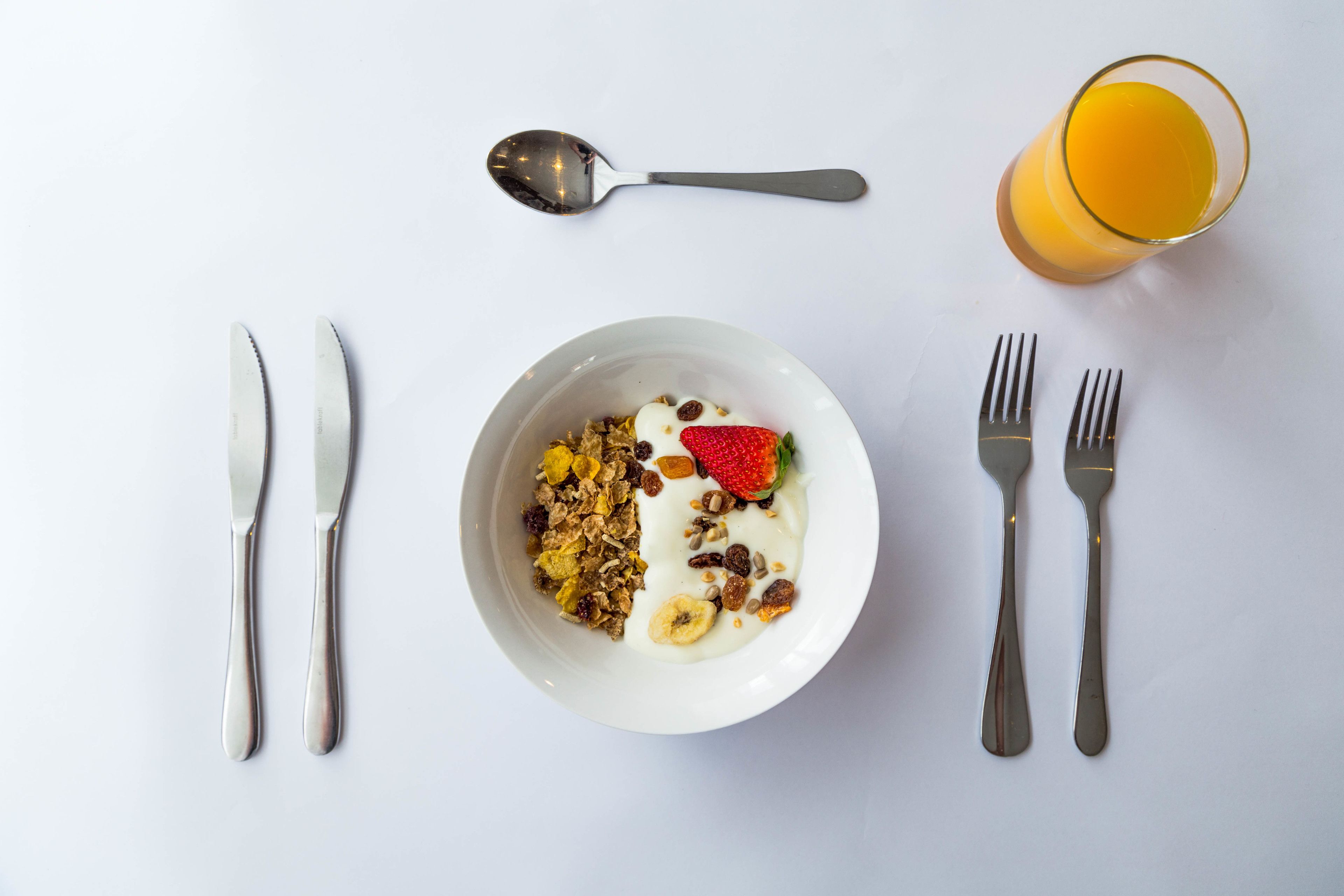 A breakfast setting with a bowl of cereal, yogurt, and fruit, accompanied by a glass of orange juice, a spoon, and cutlery on a white table.