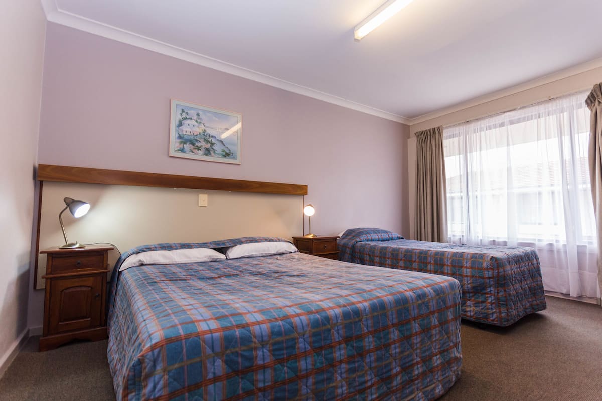 A hotel room with two beds, a wooden headboard, a nightstand with a lamp, a painting on the wall, and a window with curtains.
