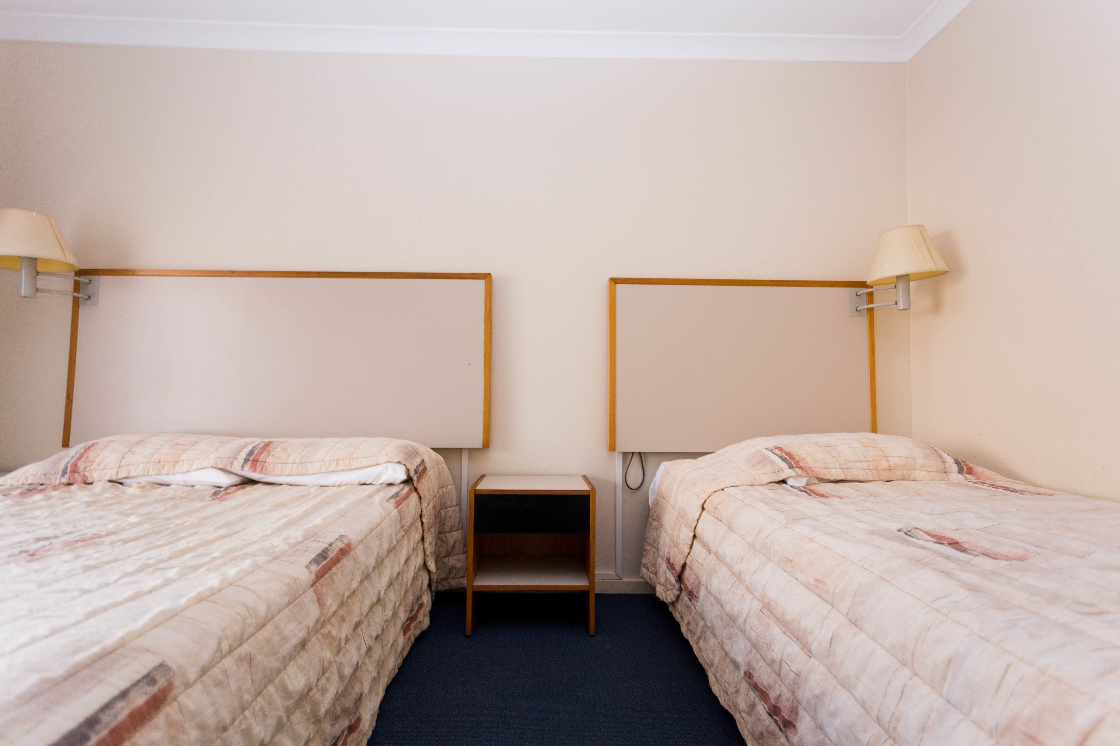 A hotel room with two single beds, each with a headboard and bedside table, and two wall-mounted lamps.