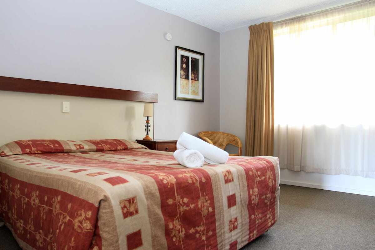 A hotel room with a large bed, a nightstand, a chair, a framed picture on the wall, and a window with curtains.