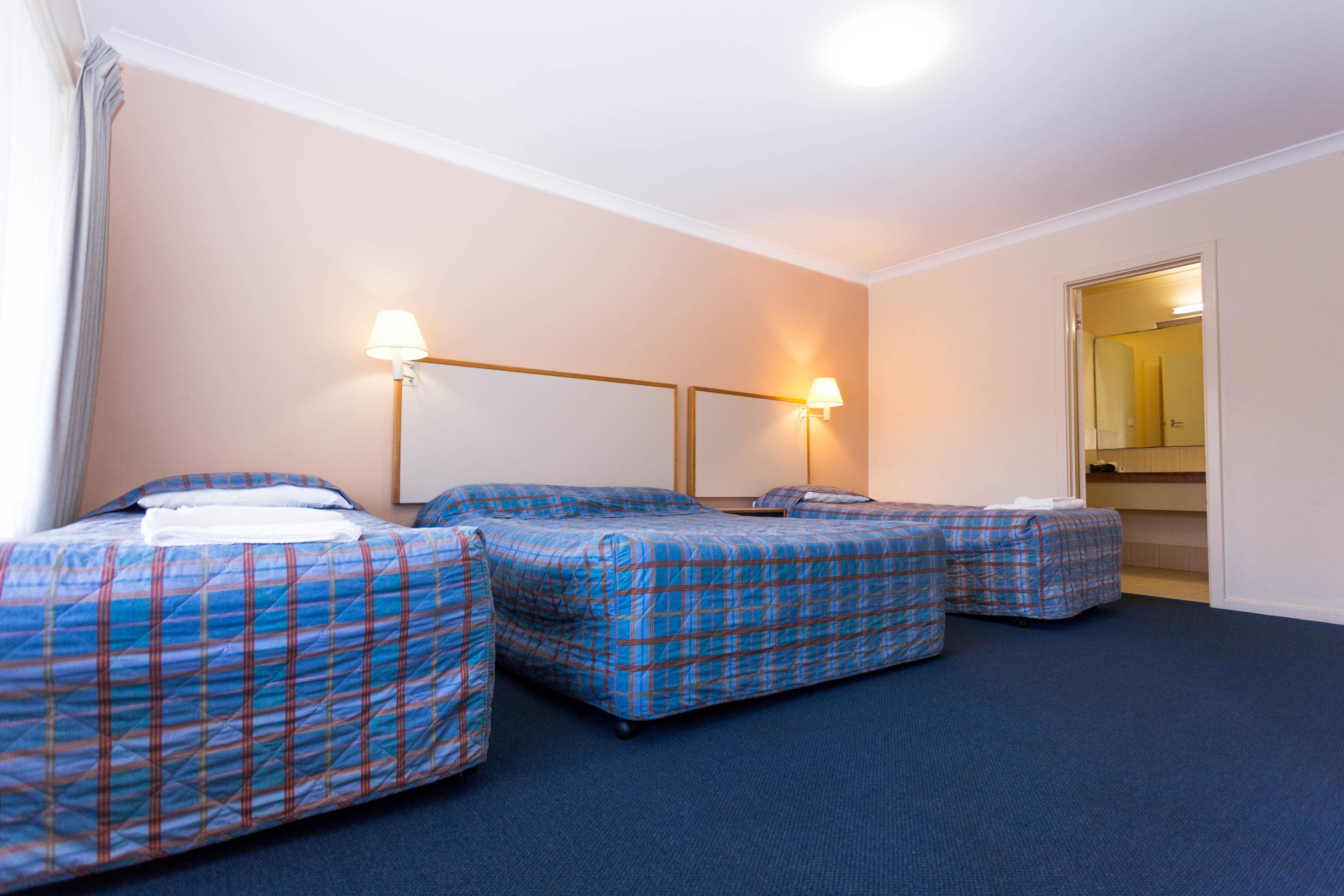 A hotel room with three beds, each with a blue plaid bedspread, a nightstand, and a wall-mounted lamp.