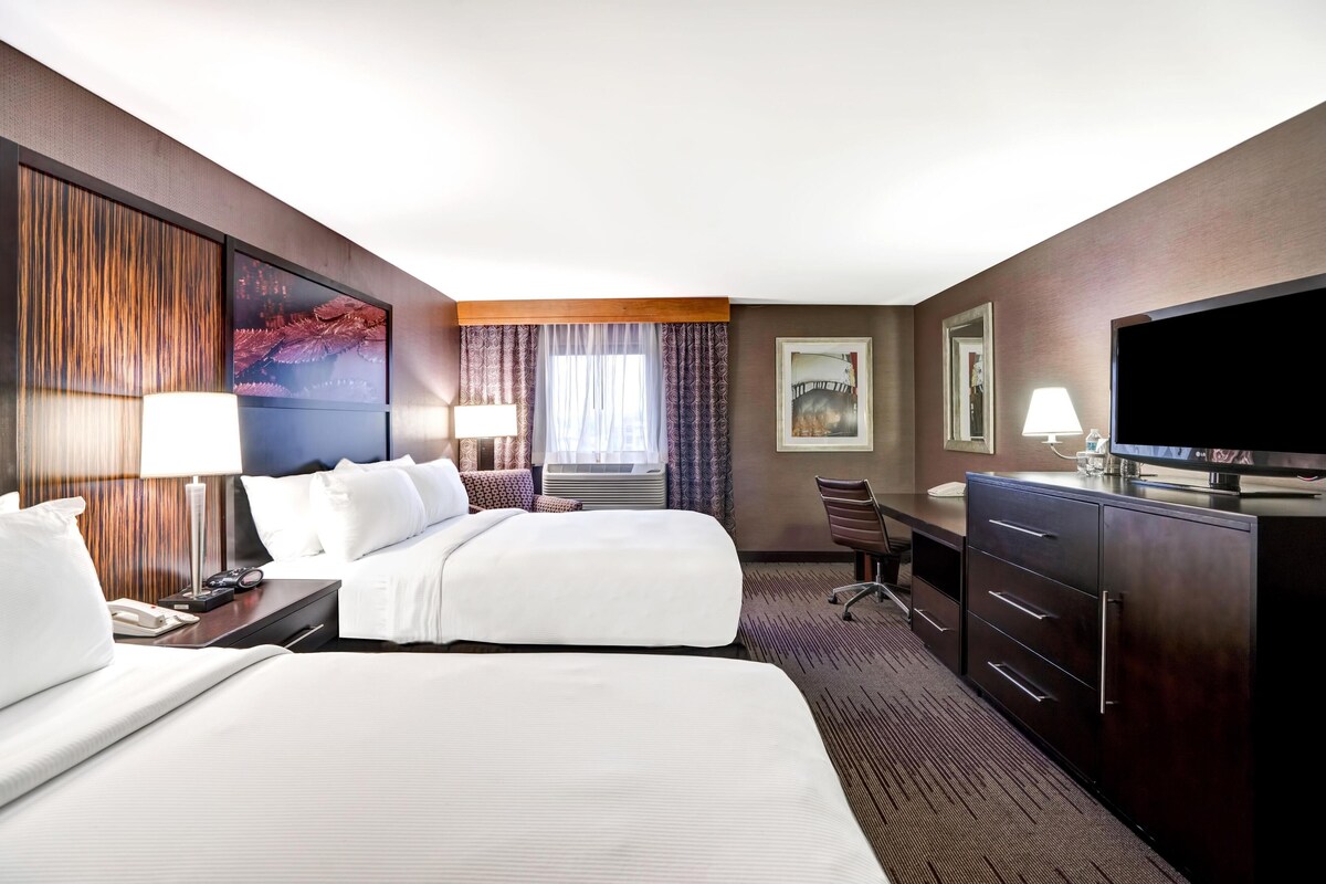 A hotel room with two beds, a desk with a lamp, a TV, and a large painting on the wall.