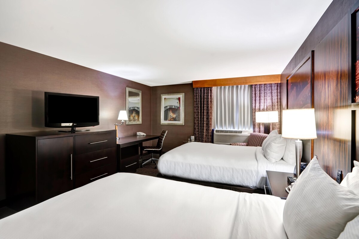 A hotel room with two beds, a desk, a television, and a window with curtains.
