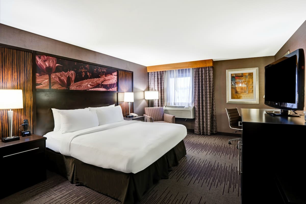A hotel room with a large bed, two bedside tables with lamps, a desk with a computer, a TV, and a large window with curtains.