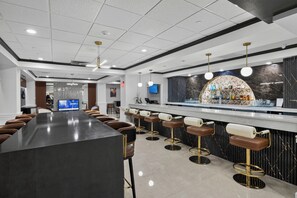 A modern bar area with a long counter, multiple stools, and a television screen in the background.