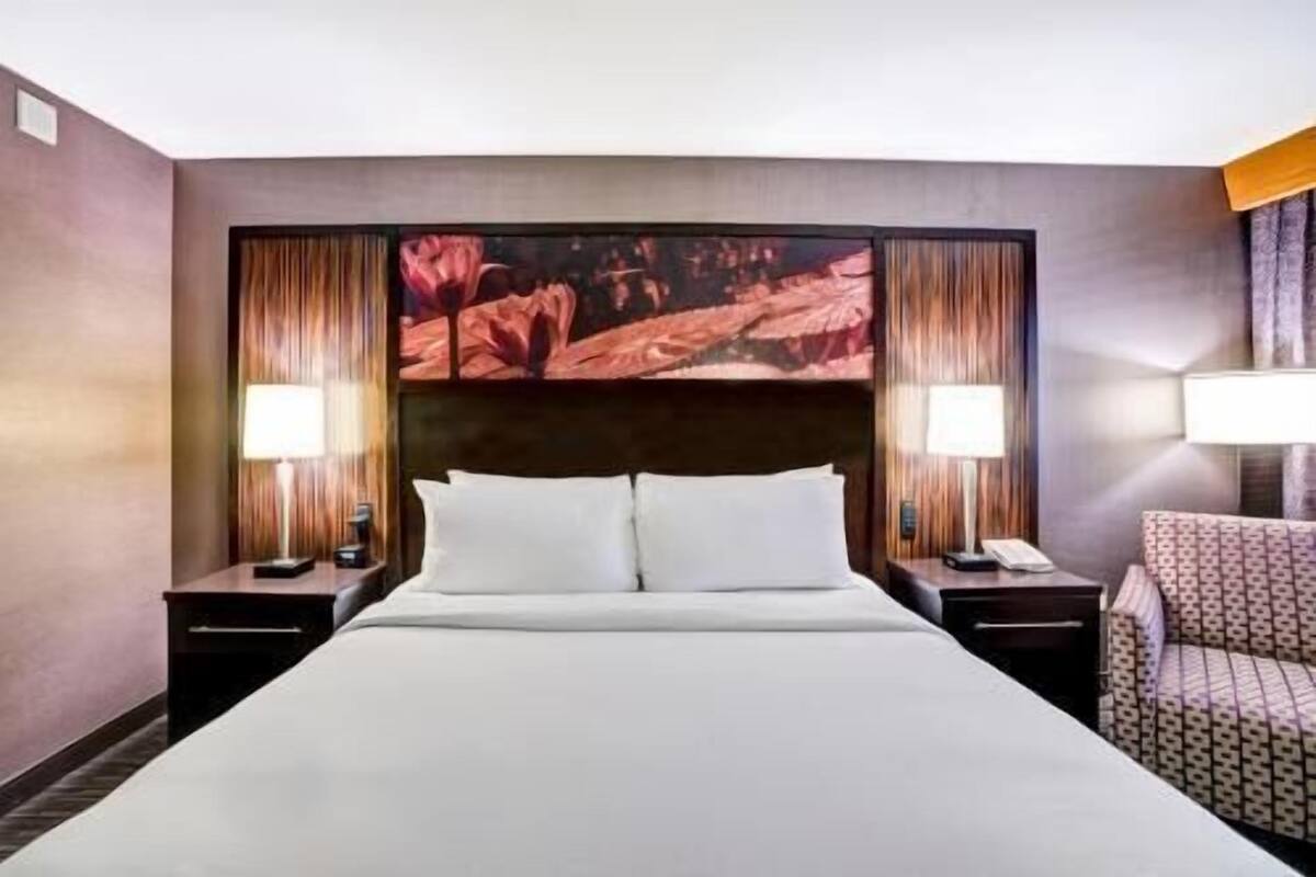 A hotel room with a large bed, two bedside tables with lamps, a chair, and a large wall mural.