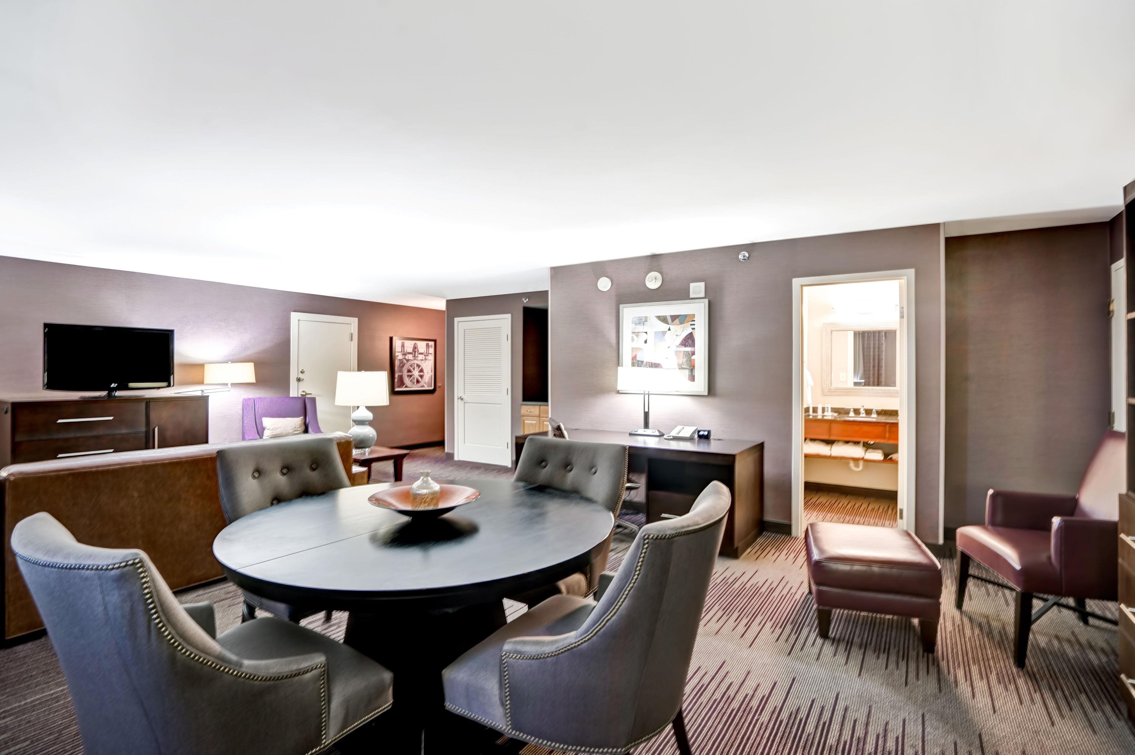 A hotel room with a round dining table, four upholstered chairs, a flat-screen TV, a desk with a lamp, and a closet with a mirror.