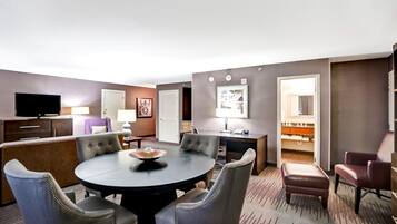 A hotel room with a round dining table, four upholstered chairs, a flat-screen TV, a desk with a lamp, and a closet with a mirror.