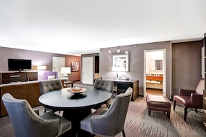 A hotel room with a round dining table, four upholstered chairs, a flat-screen TV, a desk with a lamp, and a closet with a mirror.