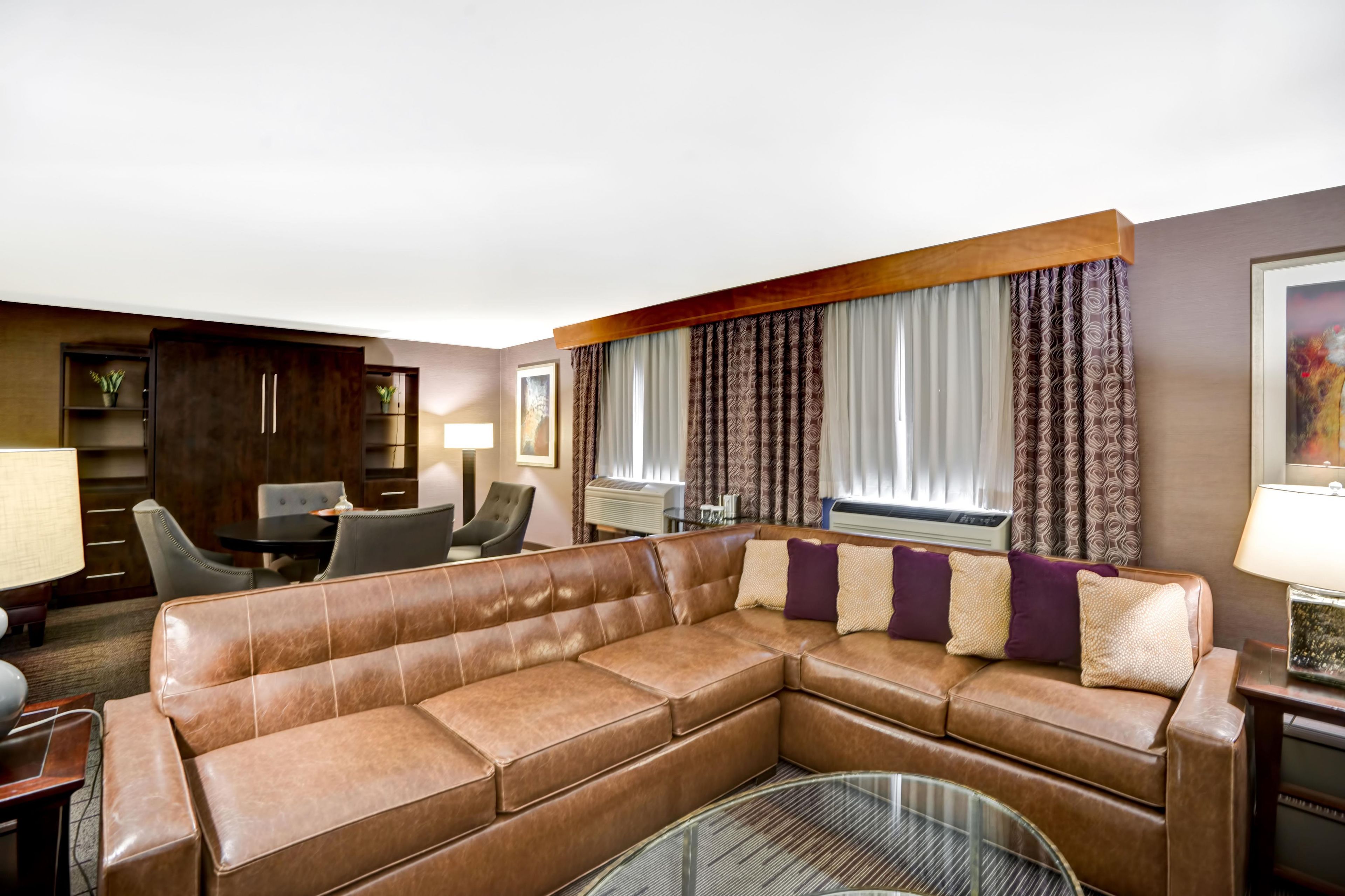 A modern living room with a leather sectional sofa, a dining area with a table and chairs, and a large window with patterned curtains.